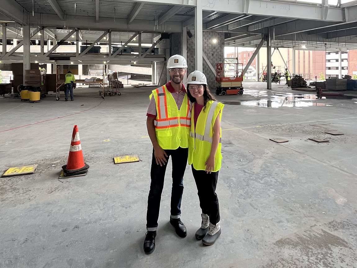 Two young people in construction helmets and vests pose for a picture at a job site